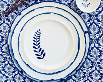Può includere: Tre piatti in ceramica bianca con bordo blu e un motivo a foglia blu. I piatti sono impilati uno sopra l'altro e sono su una tovaglia a motivi blu e bianchi.