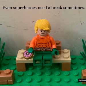 May include: A Lego minifigure of Aquaman sitting on a bench with a pink donut in front of him. The bench is made of Lego bricks and is surrounded by green grass and brown Lego bricks. The text "Even superheroes need a break sometimes." is written at the top of the image.