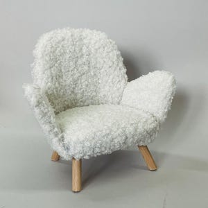 May include: A small, white, fluffy armchair with a rounded back and armrests. The chair has a soft, textured appearance and sits on four wooden legs. The overall design is minimalist and cozy, suitable for a dollhouse or miniature setting.