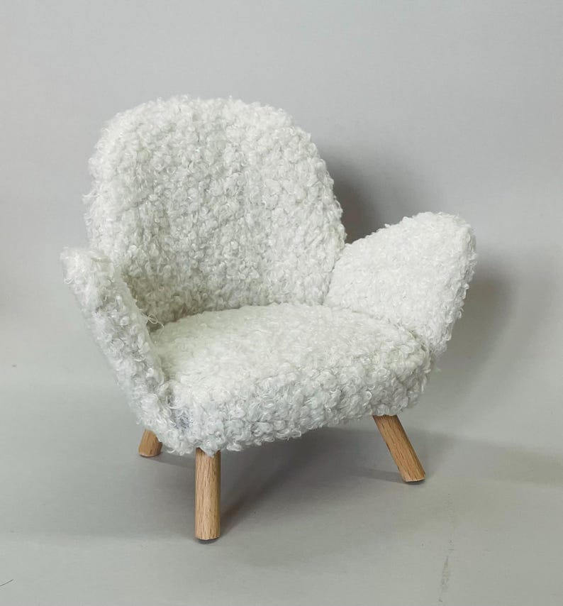May include: A small, white, fluffy armchair with a rounded back and armrests. The chair has a soft, textured appearance and sits on four wooden legs. The overall design is minimalist and cozy, suitable for a dollhouse or miniature setting.