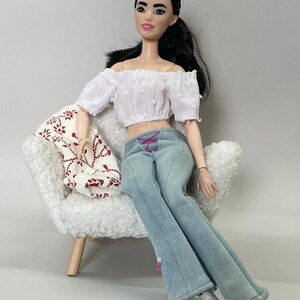 May include: A doll with black hair wearing a white off-the-shoulder crop top and light blue jeans with pink lacing. The doll is seated in a white chair with a patterned throw. The doll is wearing white sneakers.