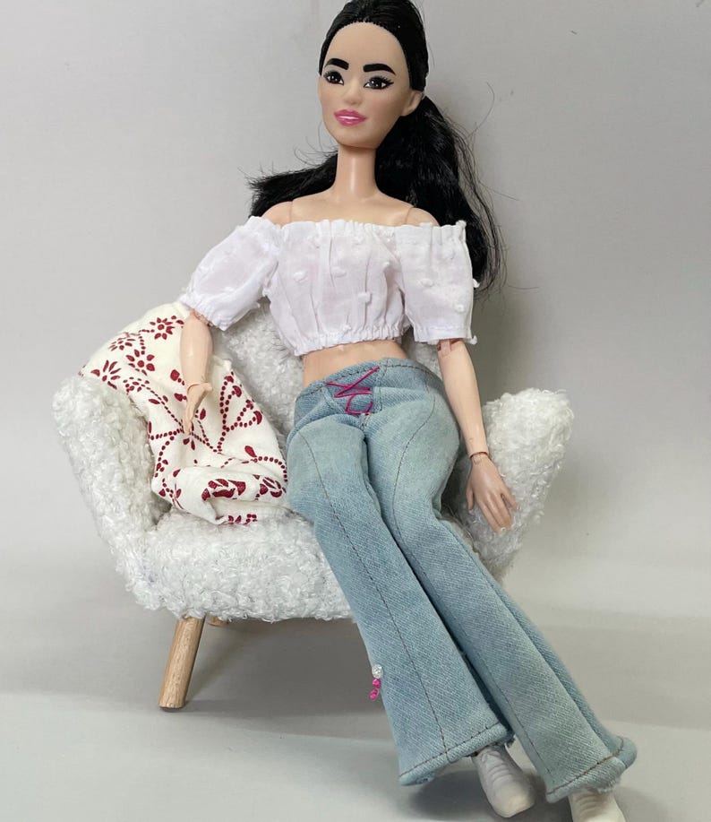 May include: A doll with black hair wearing a white off-the-shoulder crop top and light blue jeans with pink lacing. The doll is seated in a white chair with a patterned throw. The doll is wearing white sneakers.