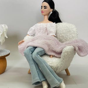 May include: A doll wearing a white off-the-shoulder top, light blue jeans, and a pink fuzzy shawl, seated in a white fluffy armchair. The doll has black hair and is posed in a relaxed position.