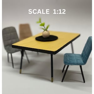 Op de afbeelding: Een miniatuur eettafelset met een gele tafel, zwarte poten en drie stoelen. De stoelen zijn grijs, beige en blauw. Een kleine plant in een witte vaas staat op de tafel.