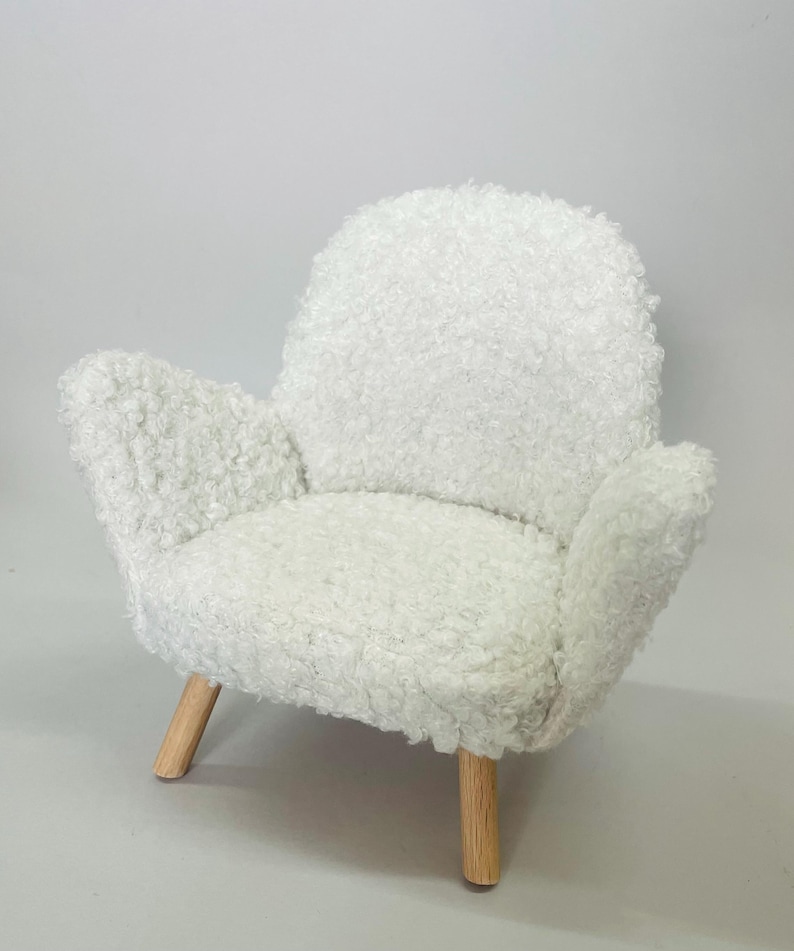 May include: A small, white, fluffy armchair with a rounded back and armrests. The chair has a soft, textured appearance and is supported by four wooden legs.