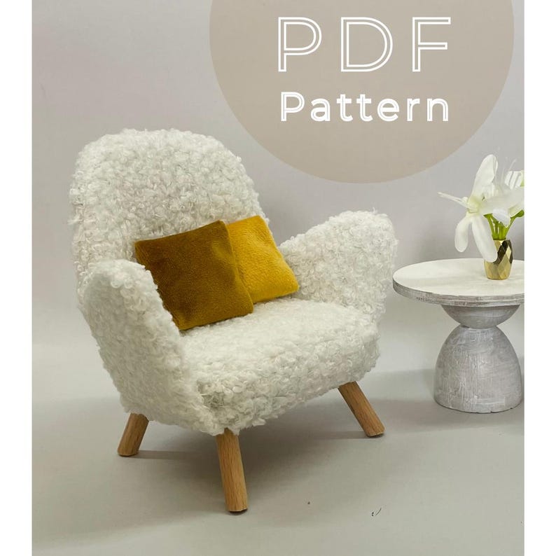 May include: A miniature white fluffy armchair with wooden legs and two square pillows in mustard and gold. A small side table with a vase of white flowers is to the right. The text "PDF Pattern" is in a circle above the chair.