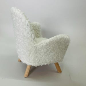 May include: A miniature white, fluffy armchair with a rounded back and armrests. The chair has light-colored wooden legs. The textured fabric gives the chair a soft, inviting appearance, perfect for a dollhouse or miniature setting.