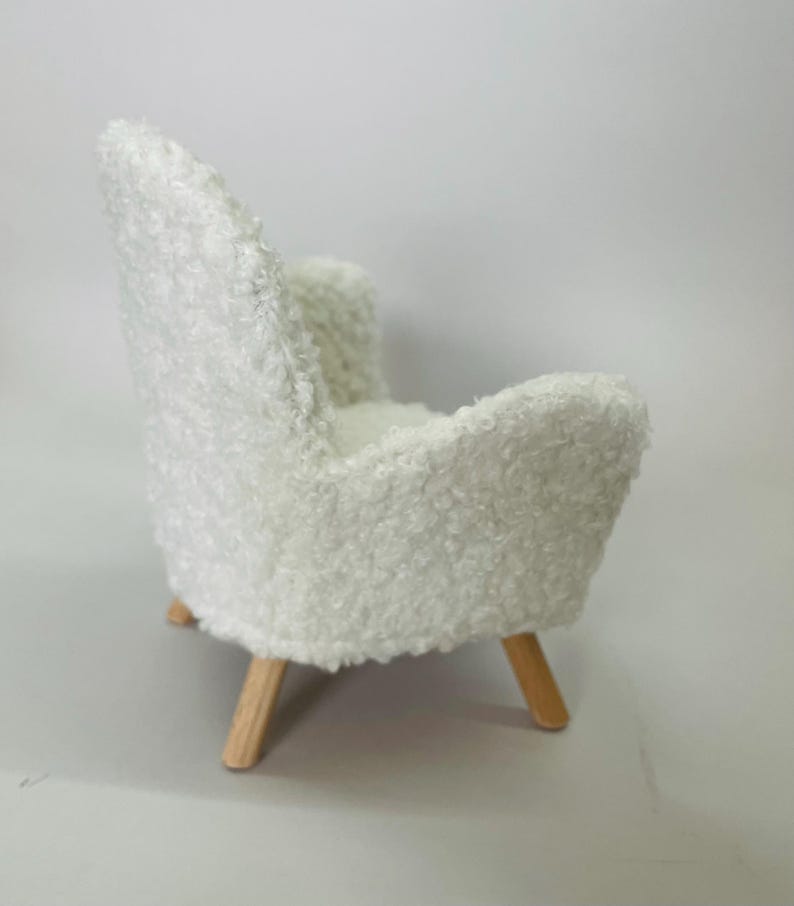 May include: A miniature white, fluffy armchair with a rounded back and armrests. The chair has light-colored wooden legs. The textured fabric gives the chair a soft, inviting appearance, perfect for a dollhouse or miniature setting.