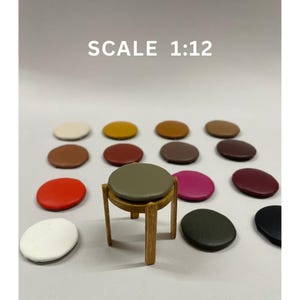 Może przedstawiać: Miniaturowy drewniany stołek z okrągłym, oliwkowozielonym siedziskiem otoczony jest kolorowymi, okrągłymi poduszkami. Tekst "SCALE 1:12" jest wyświetlany nad stołkiem. Poduszki są w różnych kolorach, w tym białym, żółtym, brązowym i czerwonym.