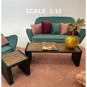 Può includere: Un divano in velluto turchese con quattro cuscini, due rosa e due bordeaux, è posizionato su un tappeto di pelo rosa. Un tavolino da caffè in legno con un cuscino a motivi floreali si trova davanti al divano. Un tavolino laterale in legno più piccolo si trova a sinistra del divano. Un vaso dorato con una pianta verde è posizionato sul tavolino da caffè. Il testo "SCALE 1:12" è stampato sopra il divano.