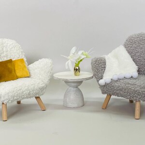May include: Miniature living room set with two plush armchairs, a small side table, and decorative accents. One white chair has two gold pillows. The other gray chair has a white blanket with pom-poms. A small table holds a gold vase with white flowers.