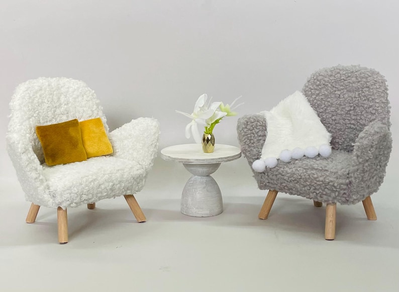 May include: Miniature living room set with two plush armchairs, a small side table, and decorative accents. One white chair has two gold pillows. The other gray chair has a white blanket with pom-poms. A small table holds a gold vase with white flowers.