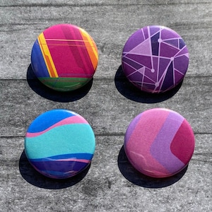 May include: Four round button pins with abstract geometric designs. The pins feature a variety of colors, including pink, purple, blue, and yellow.
