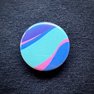 May include: A round button with a swirling pattern of blue, purple, and pink colors. The button has a glossy finish.