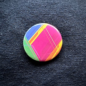 May include: A round button with a geometric design featuring a pink square with yellow, orange, blue, and green lines.