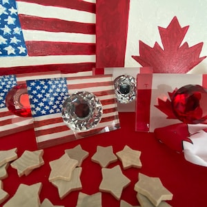 Op de afbeelding: Decoratieve glazen kastknoppen met kristal- en rode accenten, met Amerikaanse en Canadese vlagontwerpen. Houten sterren zijn verspreid over een rode ondergrond. De knoppen zijn ontworpen voor meubels.