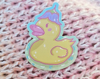 May include: A yellow rubber duck with a purple and blue flower on its head. The duck is in a blue and green bathtub with white bubbles.