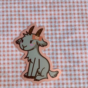 May include: A gray goat with a pink flower crown sits on a pink and white gingham background. The goat has a pink nose and a pink flower on its head.