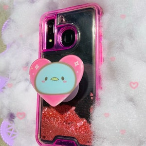May include: A pink phone case with a glitter design and a clear phone case with a blue and white penguin design. The phone case has a pop socket with a heart shape.
