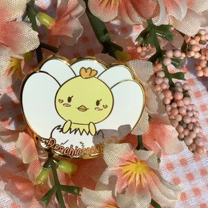 以下が含まれることがあります： 卵から孵化する漫画のひよこを描いた金色のエナメルピン。ひよこは黄色で、黒い目とピンクの頬をしています。ピンは白い花びらで囲まれており、金色の文字で「Peachicream」と書かれています。