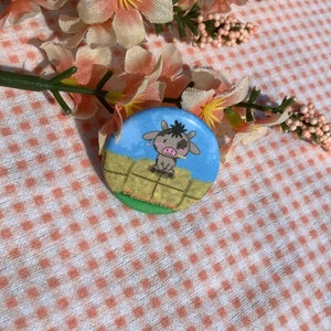Puede incluir: Un botón azul y blanco con una vaca de dibujos animados en un campo. El botón dice "Pesonacreed" en la parte inferior.