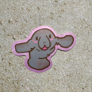 May include: A pink sticker with a cartoon illustration of a gray manatee with a pink blush on its cheeks. The manatee is smiling and has its flippers raised.