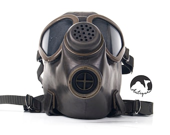May include: A vintage gas mask with a dark brown leather body and large, clear lenses. The mask has a circular air intake and adjustable straps. The lenses are framed with a bronze-colored metal.