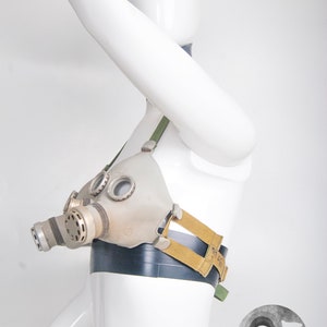 Puede incluir: Una máscara de gas vintage con una placa facial gris y plateada, una correa negra y amarilla y una correa verde. La máscara está unida a un maniquí blanco.