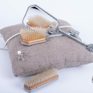 May include: Three silver-toned hairbrushes with wooden bristles and ornate handles. The brushes are arranged on a brown linen pillow with a pearl necklace and a silver spider brooch.