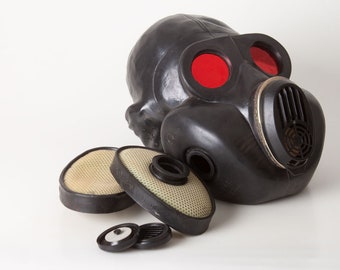 May include: A black gas mask with red eye pieces and two black filters. The mask is laying on a white surface with two additional filters and two small black pieces.