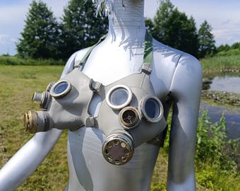 May include: A grey brassiere constructed from repurposed gas mask components, featuring circular glass lenses, metallic filter ports, and adjustable green fabric straps. The structure incorporates multiple circular apertures and brass-toned hardware.