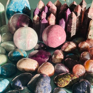 Puede incluir: Una colección de piedras y cristales pulidos de diversas formas y tamaños, incluyendo esferas, corazones y puntas. Las piedras son de tonos púrpura, azul, verde y rojo.