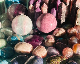 May include: A collection of polished stones and crystals in various shapes and sizes, including spheres, hearts, and points. The stones are in shades of purple, blue, green, and red.