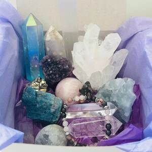 Può includere: Una scatola regalo bianca piena di vari cristalli e un braccialetto di cristallo. I cristalli includono quarzo trasparente, ametista, quarzo rosa e apatite blu. Il braccialetto è fatto di perline nere e perline di quarzo rosa.