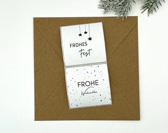 May include: A square brown envelope with two white circular stickers. The top sticker reads "FROHES Fest" with three hanging stars. The bottom sticker says "FROHE Weihnachten" with scattered dots. A sprig of frosted evergreen is in the upper right corner.