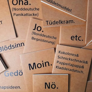 Könnte beinhalten: Eine Sammlung brauner Papierkarten mit schwarzem Text. Die Karten zeigen verschiedene deutsche Sätze und Wörter, darunter "Moin", "Nö" und "Blödsinn". Die Karten sind leicht überlappend angeordnet.