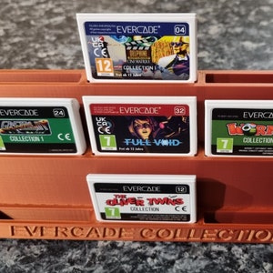 May include: A brown plastic stand with the text "EVERCADE COLLECTION" printed on the front. The stand holds five white game cartridges with colourful graphics and text, including "EVERCADE", "DELPHINE ON MAIKLE COLLECTION 1", "GREMLINS COLLECTION 1", "FULL VOID", "WORMS COLLECTION 1", and "OLIVER TWINS COLLECTION".