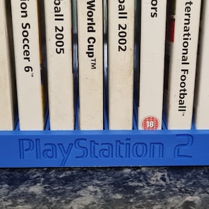 Puede incluir: Un soporte azul de plástico para juegos de PlayStation 2 con las palabras "PlayStation 2" en relieve en la parte delantera. El soporte contiene varias carcasas de juegos con lomos blancos y varios títulos de juegos.