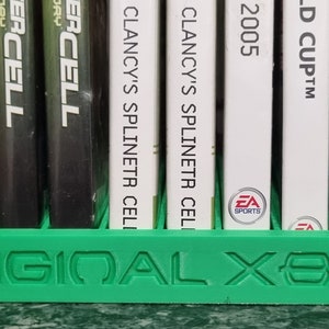 Puede incluir: Una caja de almacenamiento de plástico verde para discos de juegos de video Xbox originales. La caja tiene las palabras "ORIGINAL XBOX" en relieve en la parte delantera. La caja contiene una variedad de discos de juegos de video.