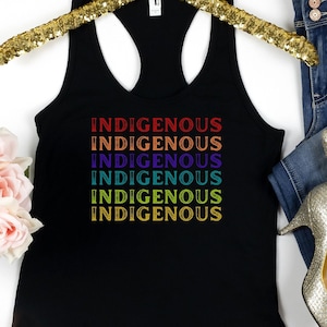 May include: Black tank top with the word "Indigenous" repeated five times in rainbow colors. The tank top is on a gold hanger and is next to a pair of blue jeans and gold heels.