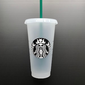 May include: A clear plastic reusable cup with a black and white sticker of a skeleton mermaid with a crown. The sticker is a parody of the Starbucks logo. The cup has a green straw.