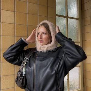Könnte beinhalten: Schwarze Lederjacke mit Reißverschluss und passendem Gürtel. Die Jacke wird mit einer hellrosa, pelzigen Kapuze kombiniert. Eine braune Leder-Umhängetasche mit dekorativem Schlüsselanhänger ist ebenfalls zu sehen.