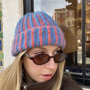 May include: A blue and coral striped knitted beanie hat. The hat is worn with brown oval sunglasses and a brown jacket. A warm accessory for cold weather.