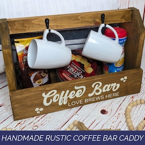 May include: A brown wooden caddy with two black metal hooks holding two white ceramic mugs. The front panel features white text that reads Coffee Bar and LOVE BREWS HERE, with small coffee bean graphics. The caddy contains food items.