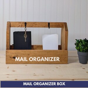 May include: A rectangular wooden mail organizer box with a medium brown finish. The front panel features the white text MAIL ORGANIZER. The top bar includes two black metal hooks, one holding a silver key on a ring. A black notebook and white papers are inside.