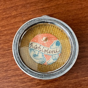 Pode incluir: Um medalhão redondo prateado com uma borda decorativa. No interior, uma imagem circular apresenta uma faixa com o texto "S. Ghisleri" e um desenho floral. O fundo é um tecido texturizado amarelo dourado.