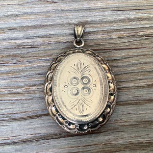 May include: A silver oval locket with a floral design and a decorative border. The locket has a loop at the top for attaching a chain.
