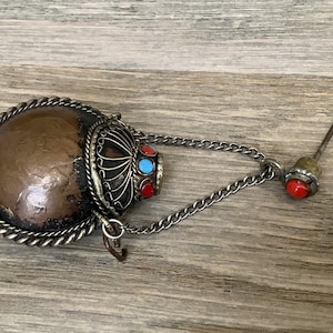 May include: An antique, copper-toned flask with a decorative silver-colored metal trim and chain. The flask features a detailed design and is adorned with red and turquoise-colored stones. A small stopper with a red stone is attached.