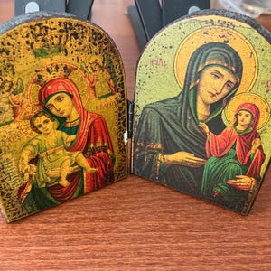 Puede incluir: Un icono de madera con bisagras que presenta imágenes religiosas. El icono representa a la Virgen María y el niño, con halos dorados y obras de arte detalladas. Los colores incluyen rojo, verde, dorado y negro, con una apariencia envejecida.