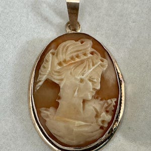 Peut inclure: Pendentif camée ovale représentant un profil de femme sculpté, serti dans un cadre doré. Le camée présente des détails complexes de la coiffure et des vêtements de la femme. Il possède une bélière dorée pour un collier.
